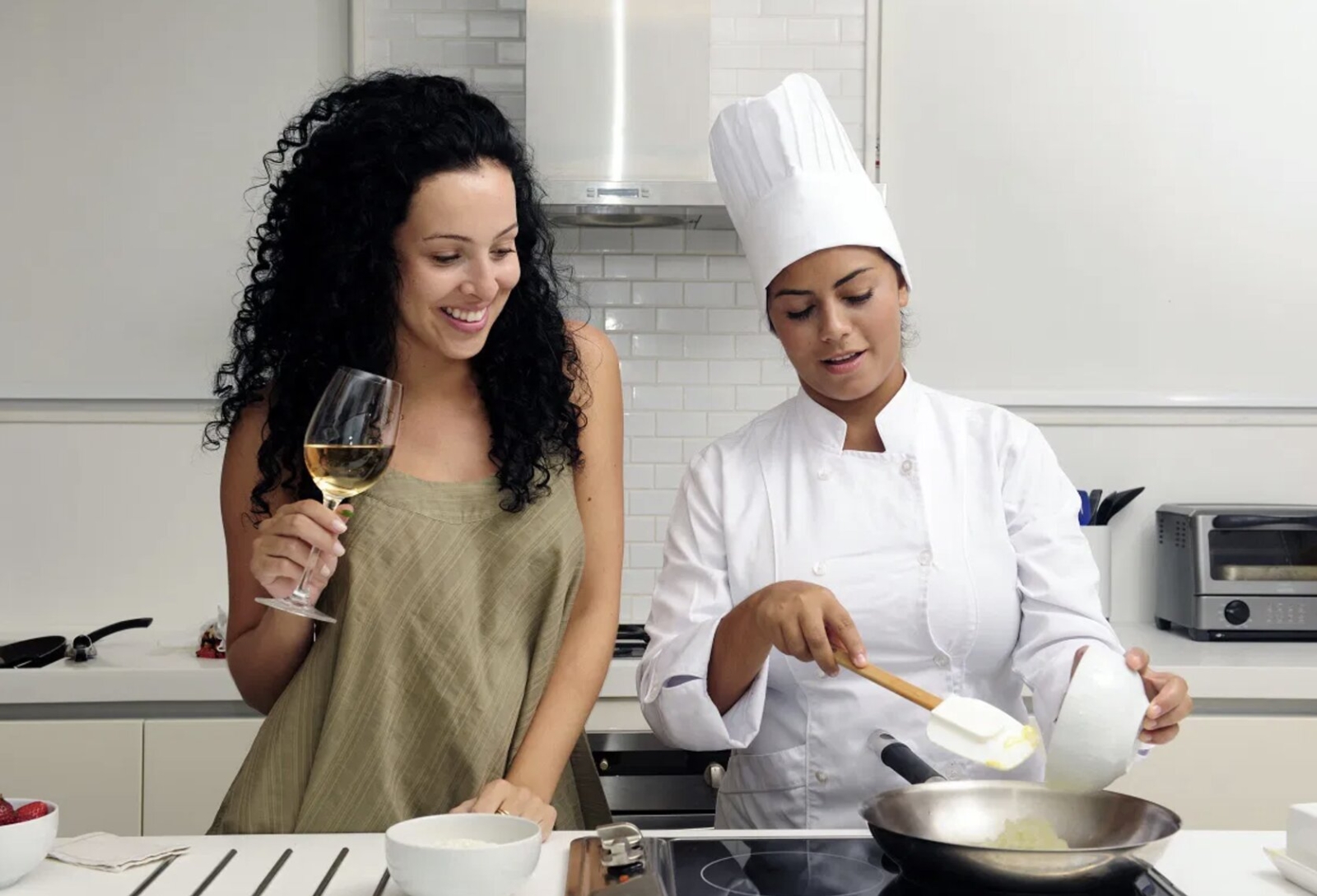 A woman smiling while participating in a cooking class, watching a professional chef prepare food in a modern kitchen.
